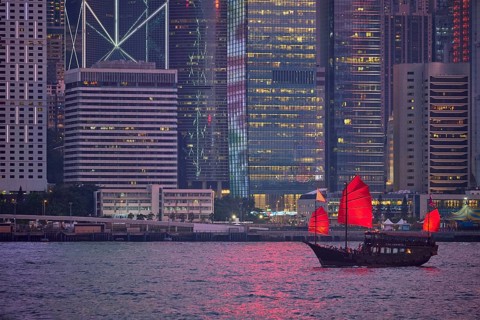 Tourist junk boat ferry with red sails and Hong Kong skyline cityscape downtown skyscrapers over Victoria Harbour in the evening. Hong Kong