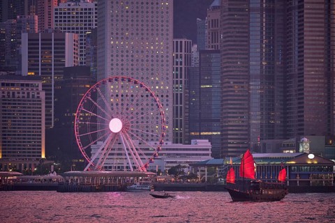 Tourist junk boat ferry with red sails and Hong Kong skyline cityscape downtown skyscrapers over Victoria Harbour in the evening. Hong Kong