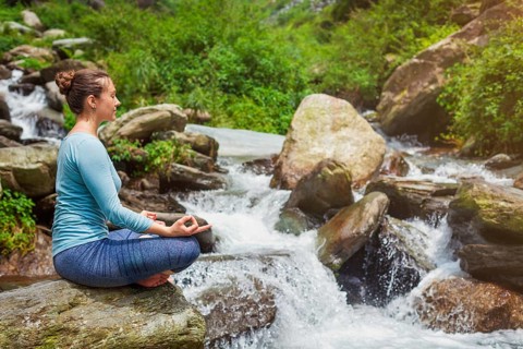 Woman in Hatha yoga asana Padmasana outdoors at tropical waterfall