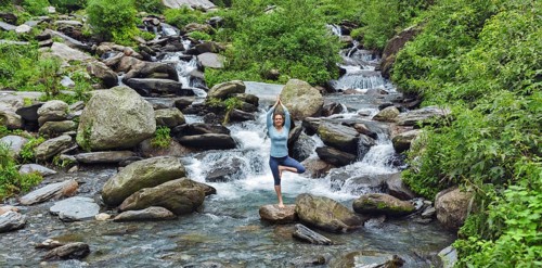 Woman practices balance yoga asana Vrikshasana tree pose at waterfall outdoors. Himachal Pradesh