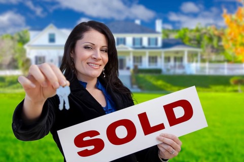 Hispanic woman holding sold sign and keys in front of beautiful house