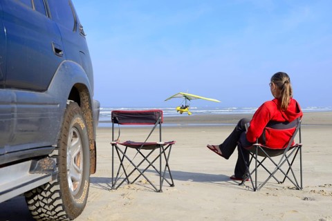 Woman sitting on camping chair next to all-terrain vehicle on beach watching a light aircraft taking off
