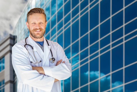 Handsome young adult male doctor with beard in front of hospital building