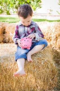 Cute young mixed-race boy sitting on hay bale putting coins into pink piggy bank
