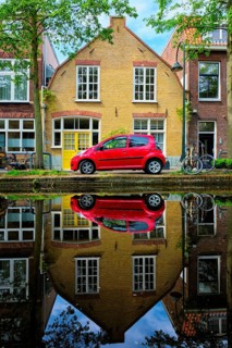 Red car on canal embankment in street of Delft with reflection and bicycle