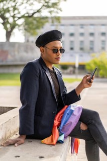 Latino gay male with makeup on wearing trendy hat holding a cellphone and lgbt flag in a park