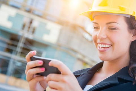 Young professional female contractor wearing hard hat at contruction site texting with cell phone