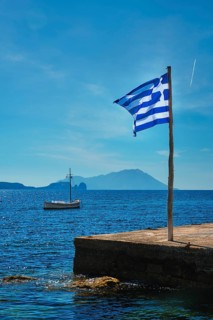 Greek flag in the blue sky on pier and traditional greek fishing boat in the Aegean sea with greek flag