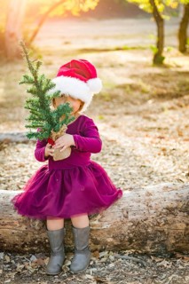 Cute mixed-race young baby girl having fun with santa hat and christmas tree outdoors on log