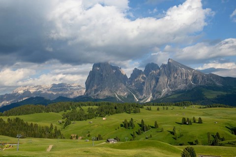 In Suedtirol auf der Seiser Alm im Fruehsommer mit einigen Almhuetten und einem Schilift auf saftig gruenen Weiden. Im Hintergrund ist ein grosser felsiger Berg mit einigen Wolken am blauem Himmel
