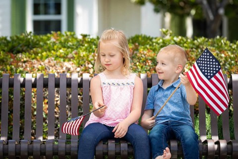 Young sister and brother comparing each others american flag size on the bench at the park