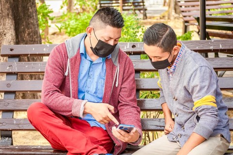 Two men wearing masks checking their cell phones on a bench