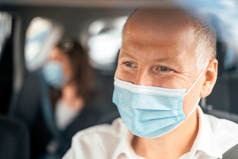 A happy taxi driver wearing a mask and the passenger on the back seat of the car in the background