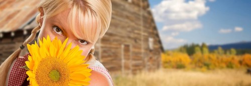 Beautiful caucasian young woman holding sunflower in front of rustic barn in the country