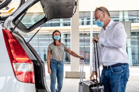A taxi or Uber driver helping a passenger in a protective mask with her luggage at the airport