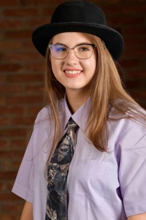 Fotostudio Portrait einer jungen selbstbewusste Frau vor einer Mauer aus roten Ziegeln. Das Maedchen traegt ein Hemd mit Krawatte und einen schwarzen Hut. Das herzhafte laecheln zeigt ihre positive Le...
