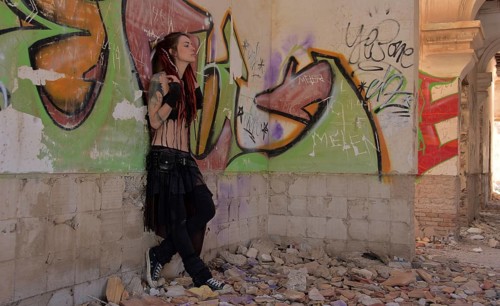 Young woman with tatoos and dreadlocks stands against wall with graffiti of an abandoned house