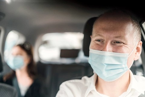A happy taxi driver wearing a mask and the passenger on the back seat of the car in the background