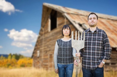 Biracial chinese and caucasian young couple holding wooden pitch fork in front of rustic barn in the country