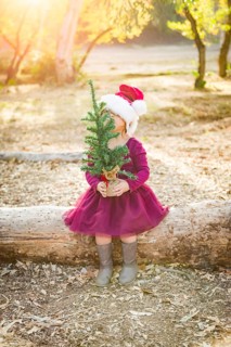 Cute mixed-race young baby girl having fun with santa hat and christmas tree outdoors on log