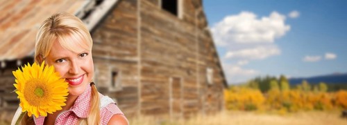 Beautiful caucasian young woman holding sunflower in front of rustic barn in the country