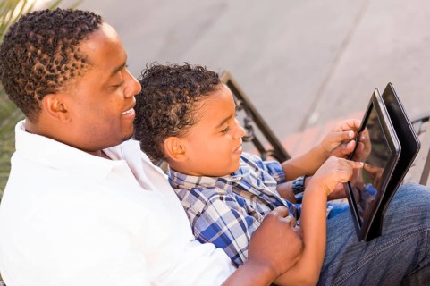 African american father and mixed-race son using computer tablet on bench in park