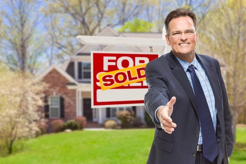 Smiling male agent reaching for a hand shake in front of beautiful house and sold real estate sign