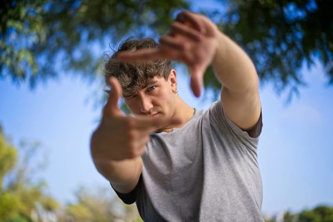 A handsome young man blinks his eyes and looks through a frame made up of hands. Attractive man make frame by fingers