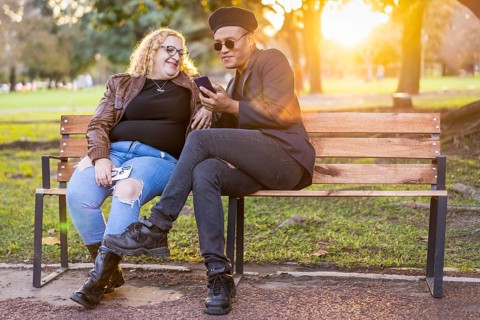 Gay friends sitting on a bench in a park at sunset chatting and looking at the cell phone