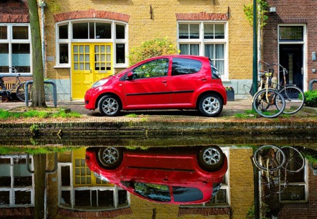 Red car on canal embankment in street of Delft with reflection and bicycle