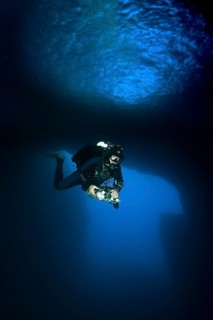 Diver dives with underwater lamp through rock grotto underwater cave Blue Dome with blue light in Blue Hole of Gozo