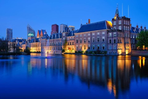 View of the Binnenhof House of Parliament and the Hofvijver lake with downtown skyscrapers in background illuminated in the evening. The Hague