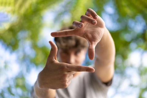 A handsome young man blinks his eyes and looks through a frame made up of hands. Attractive man make frame by fingers