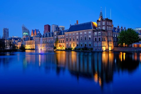 View of the Binnenhof House of Parliament and the Hofvijver lake with downtown skyscrapers in background illuminated in the evening. The Hague
