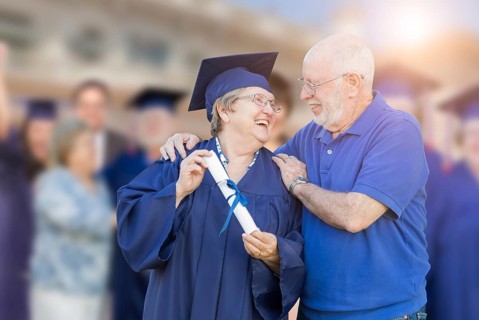 Senior woman in hat and gown being congratulated by husband at outdoor graduation ceremony