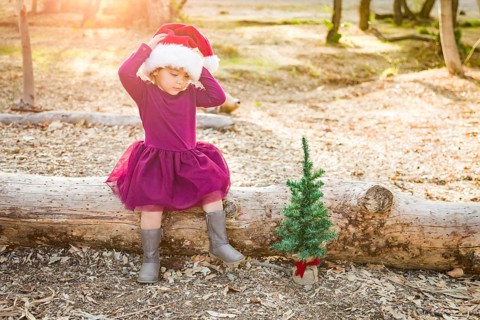 Cute mixed-race young baby girl having fun with santa hat and christmas tree outdoors on log