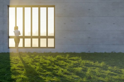 Woman standing at window with bright sun light shining through concrete wall in grass and rocks field