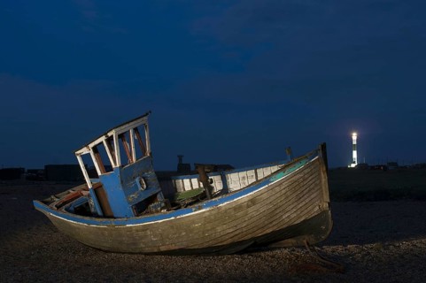 Abandoned fishing boat on shingle beach with distant lighthouse at night