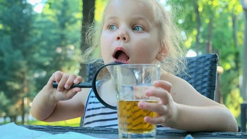 Little girl looks into the lens around. Close-up of blonde girl studying the world around her looking at it through magnifying glass while sitting in street cafe on the park