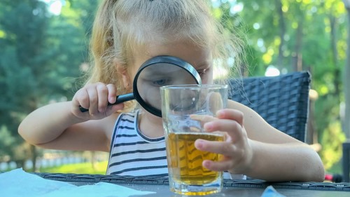 Cute little girl carefully looks into the lens at the juice in the glass. Close-up of blonde girl studying her juice in glass while looking at her through magnifying glass while sitting in street cafe...