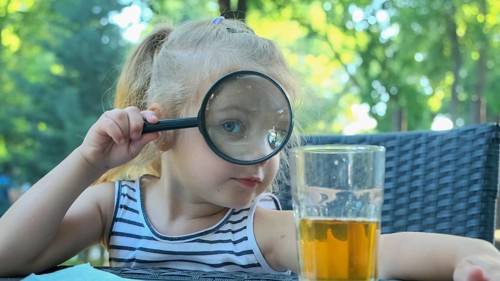 Little girl looks into the lens around. Close-up of blonde girl studying the world around her looking at it through magnifying glass while sitting in street cafe on the park. Odessa Ukraine