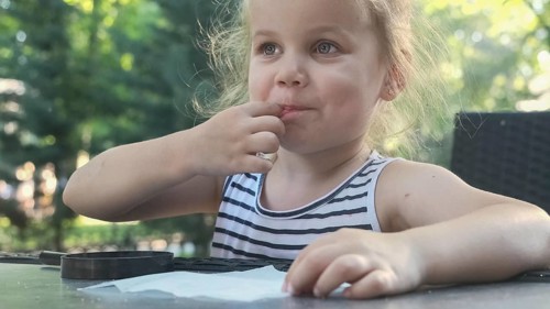 Little girl tastes salt. Close-up portrait of blonde girl takes salt from napkin with her finger and tastes it while sitting in street cafe on the park. Odessa Ukraine