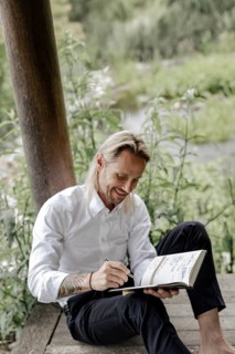 Businessman in white shirt sits on wooden jetty and writes in a book,