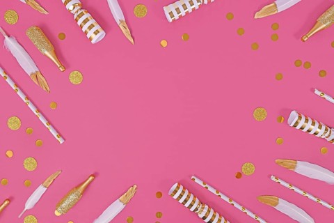 Party flat lay with golden and white decoration like confetti and feathers forming border around pink background,