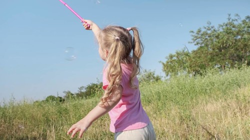 Little girl catches soap bubbles in butterfly net while playing in tall grass in park. Cute little girl smiles and catches on soap bubbles in aerial insect net in meadow on sun day. Odessa, Ukraine