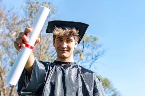 Happy graduated young man wearing a bachelor gown and a black mortarboard and showing his diploma,