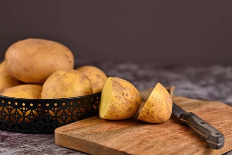 Cutting raw potato with peel on wooden cutting board with pile of whole potatoes in background,