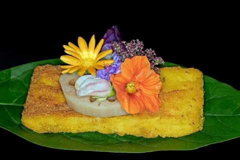Fried polenta slice with a slice of lotus root and meadow flowers as decoration on an avocado leaf, foddfotography with black background