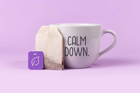 Concept for calming herbal tea with tea bag with leaf on label next to tea cup with text Calm down on violet background,