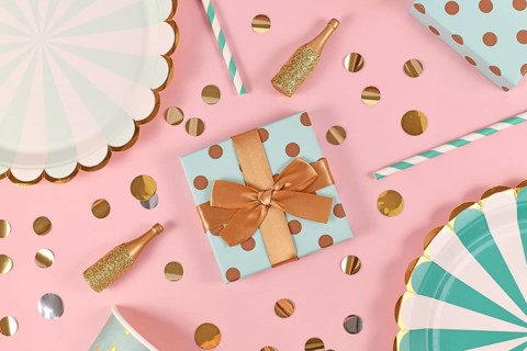 Birthday gift box with with teal blue paper plates, small golden champagne bottles and confetti on pink background